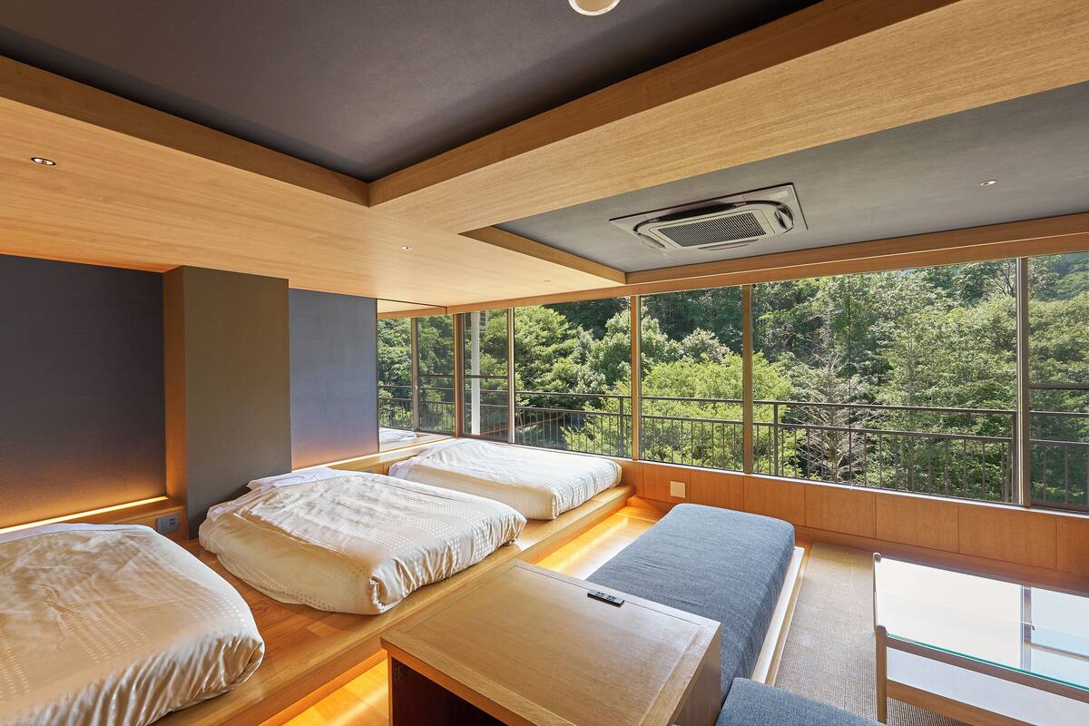 A modern room with two beds, a small table, and large windows offering a view of a forest.