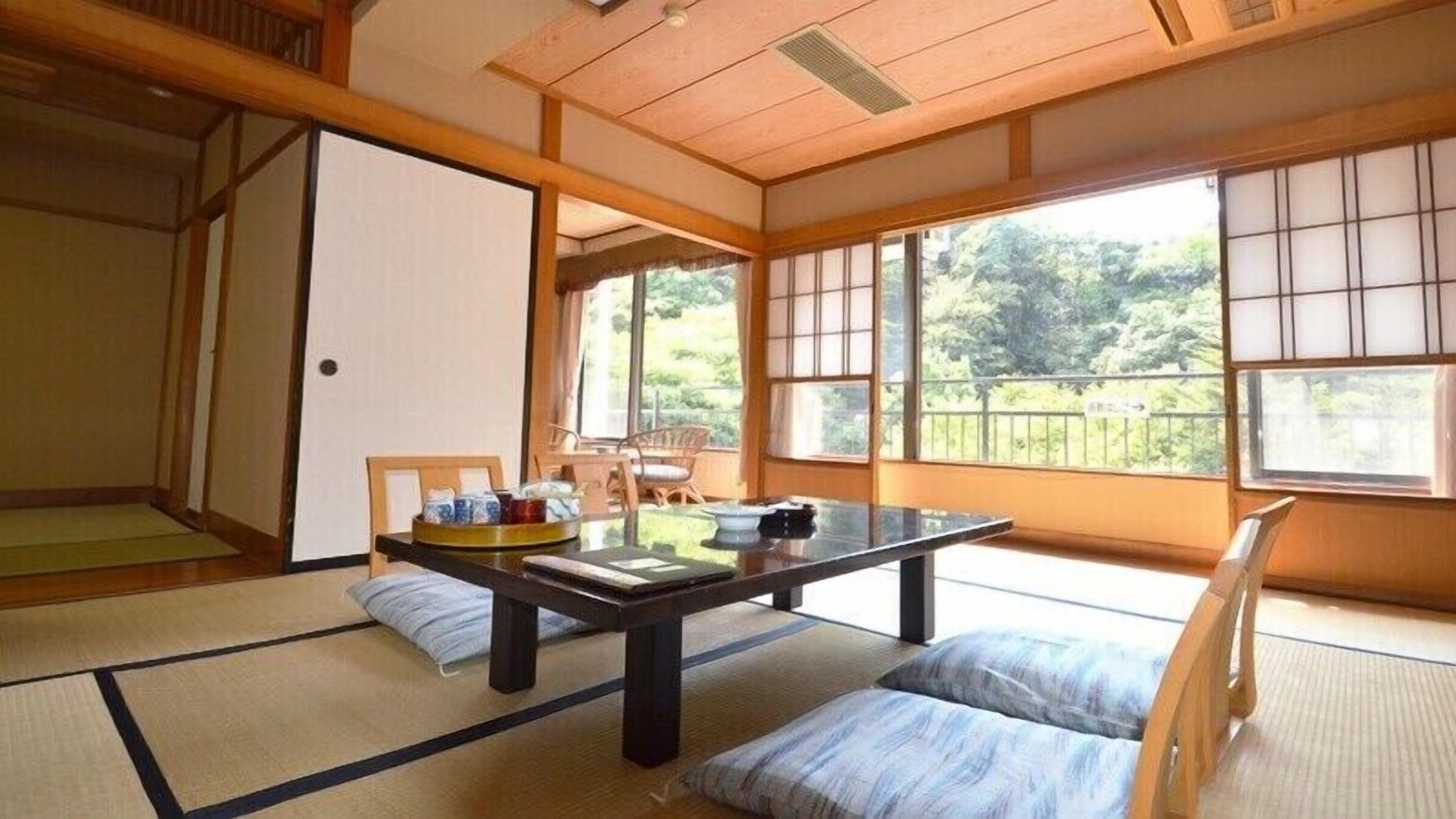 Um quarto japonês tradicional com tatames, uma mesa baixa com almofadas e amplas janelas com vista para o verde.