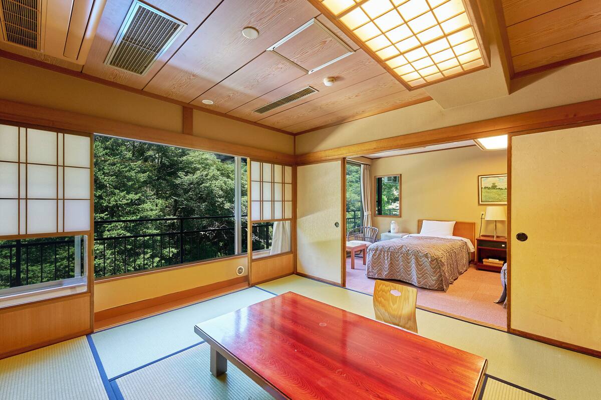 A traditional Japanese room with tatami flooring, a wooden table, and a view of a forest through large windows.