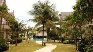 A pathway leading to a building with a pool, surrounded by palm trees and greenery. - Talay Samran by Lease Back Thailand (Cha-am)
