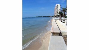 A beach with a concrete walkway, palm trees, and a tall building in the background. - Talay Samran by Lease Back Thailand (Cha-am)