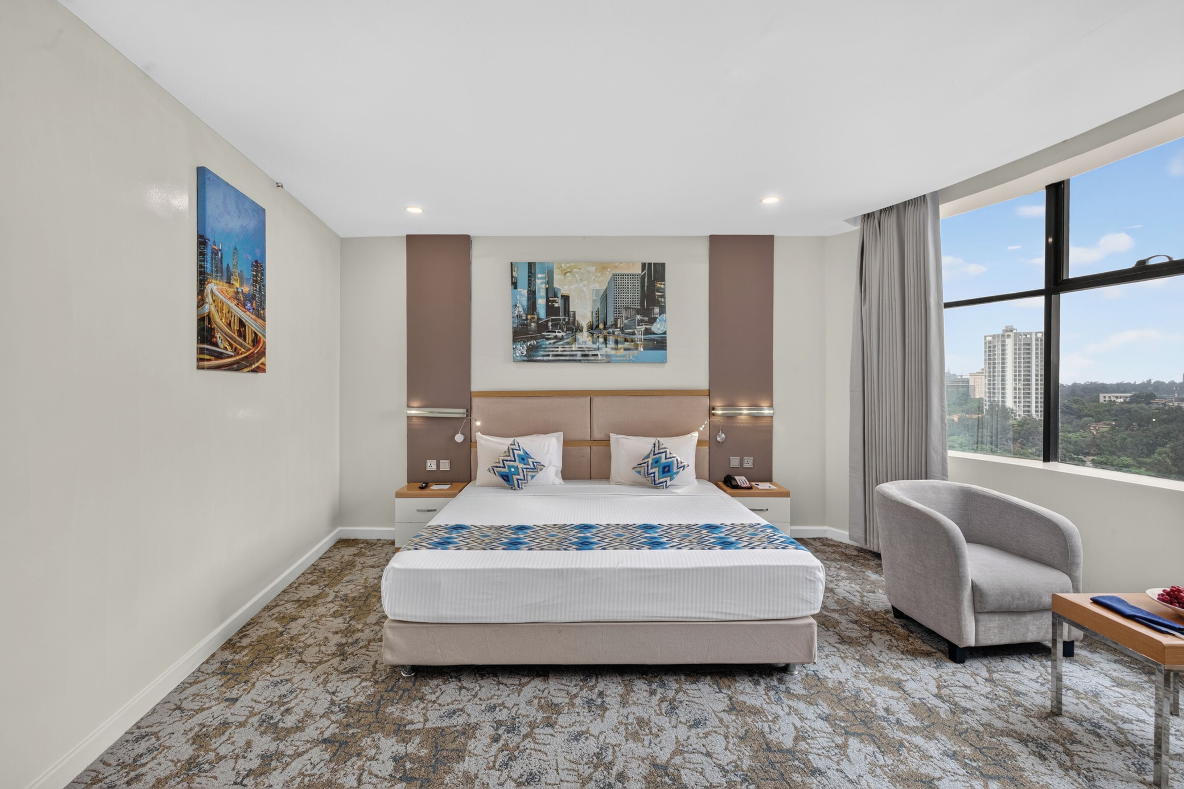 Une chambre d’hôtel avec un grand lit, deux tables de chevet, une chaise, une fenêtre donnant sur la ville et des œuvres d’art accrochées aux murs.