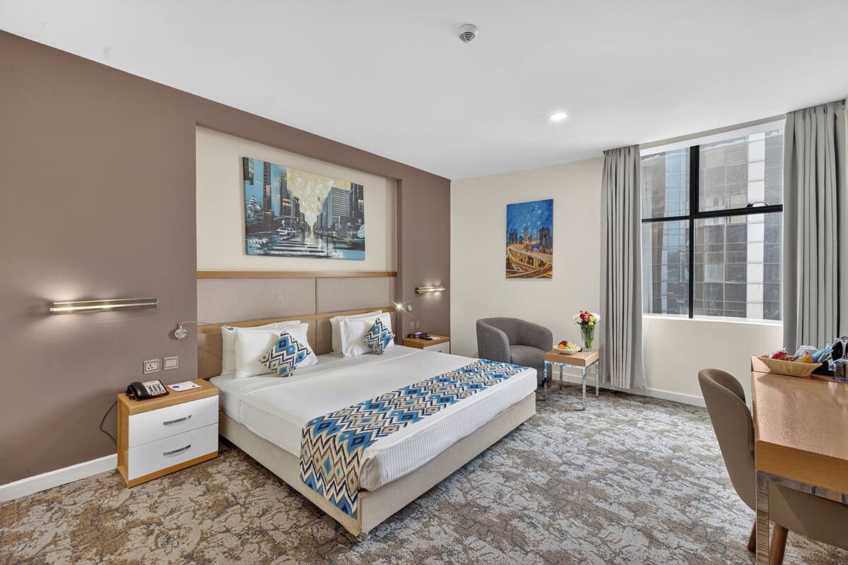 A hotel room with a bed, bedside tables, a desk, a chair, and artwork on the walls.