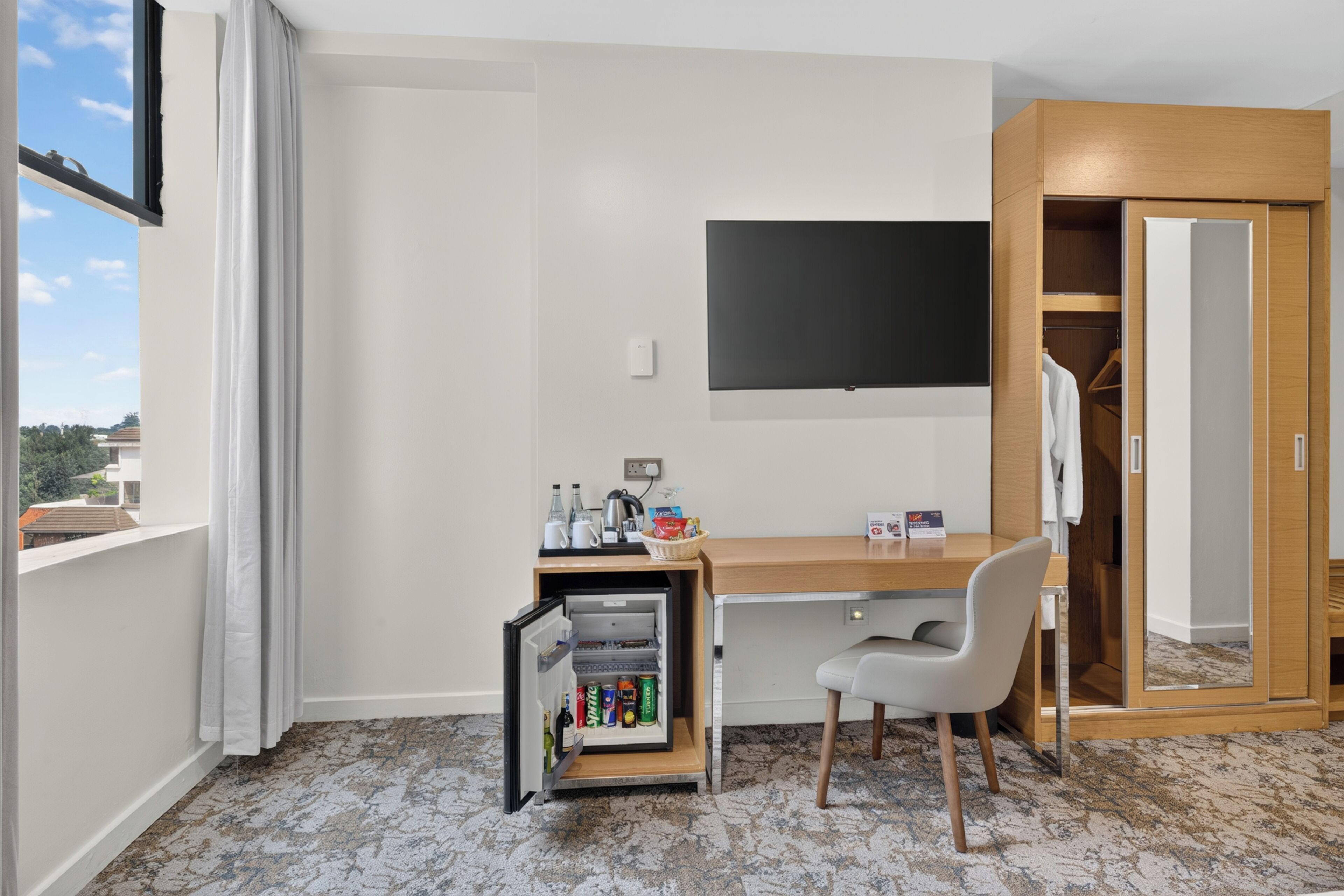Una camera d'albergo con scrivania, mini-frigorifero, televisore, armadio e finestra con tende.