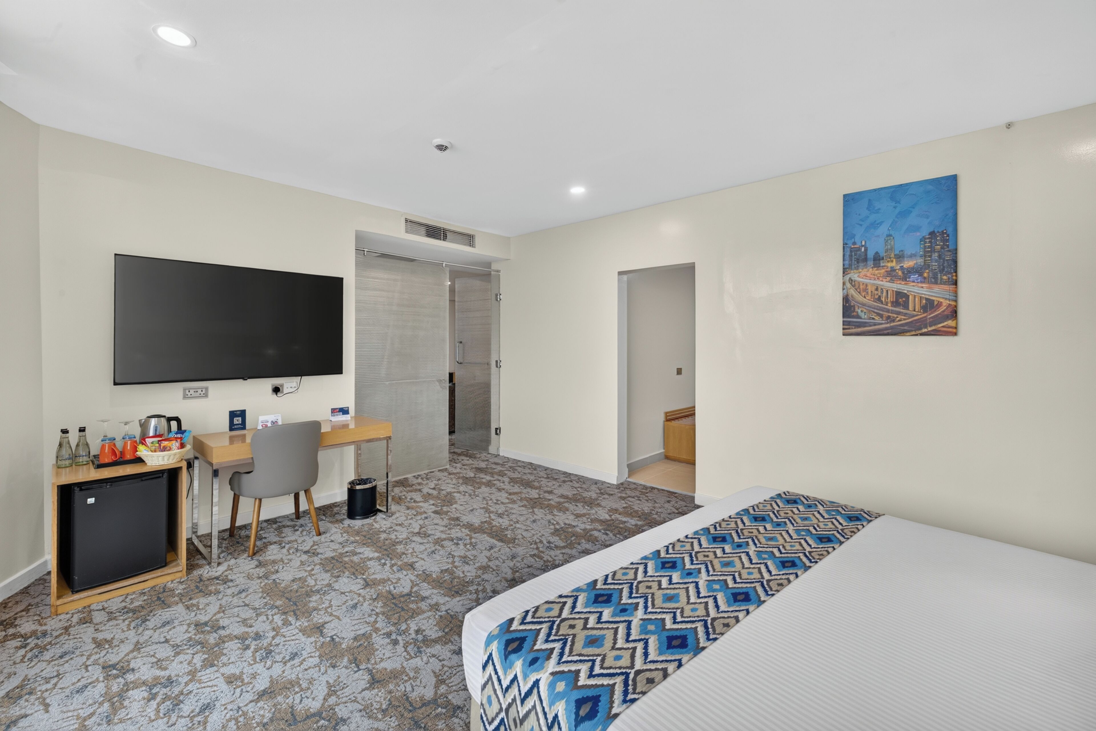 A hotel room with a bed, desk, chair, TV, and a painting on the wall.
