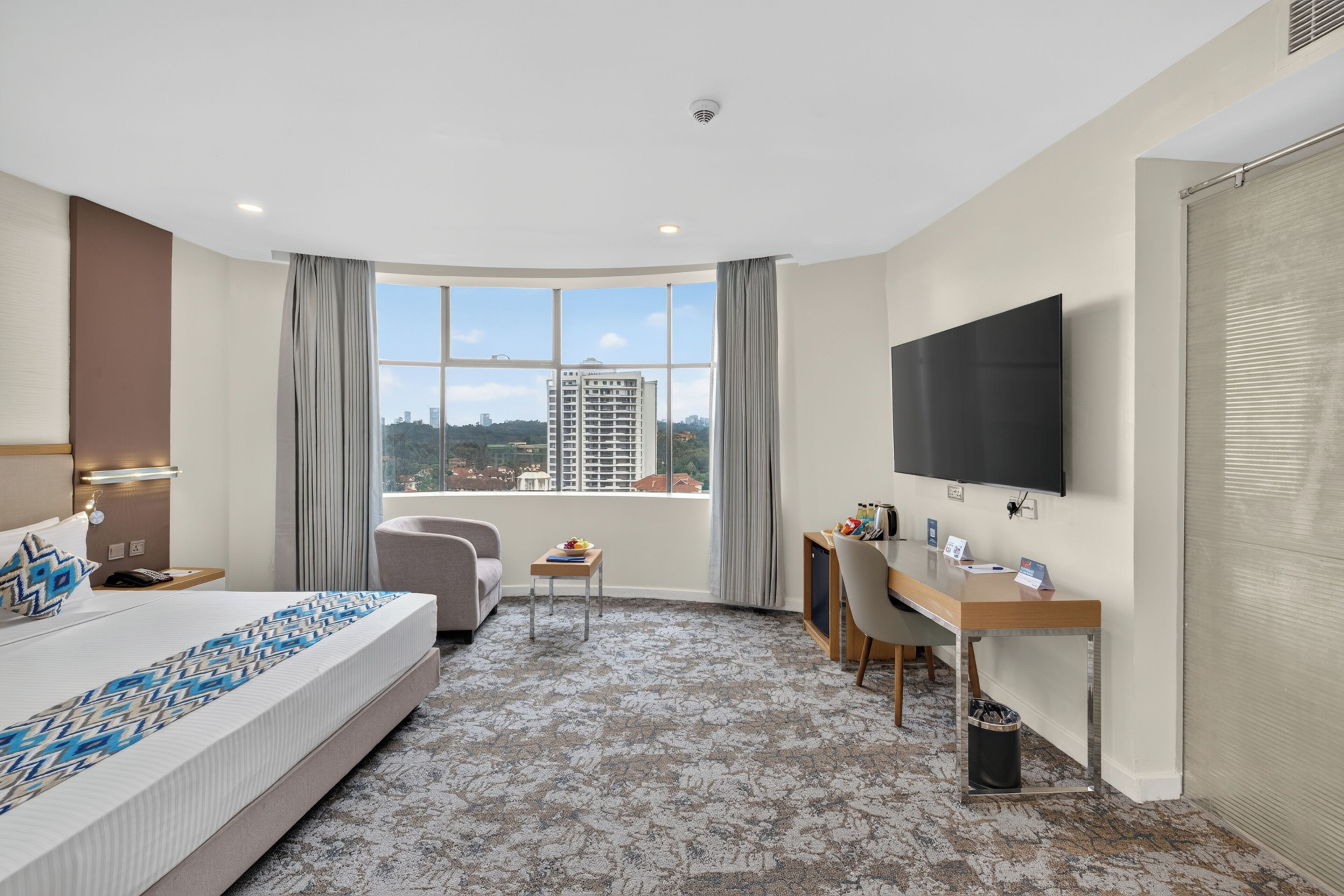 A hotel room with a bed, a desk, a chair, a TV, and a view of the city.