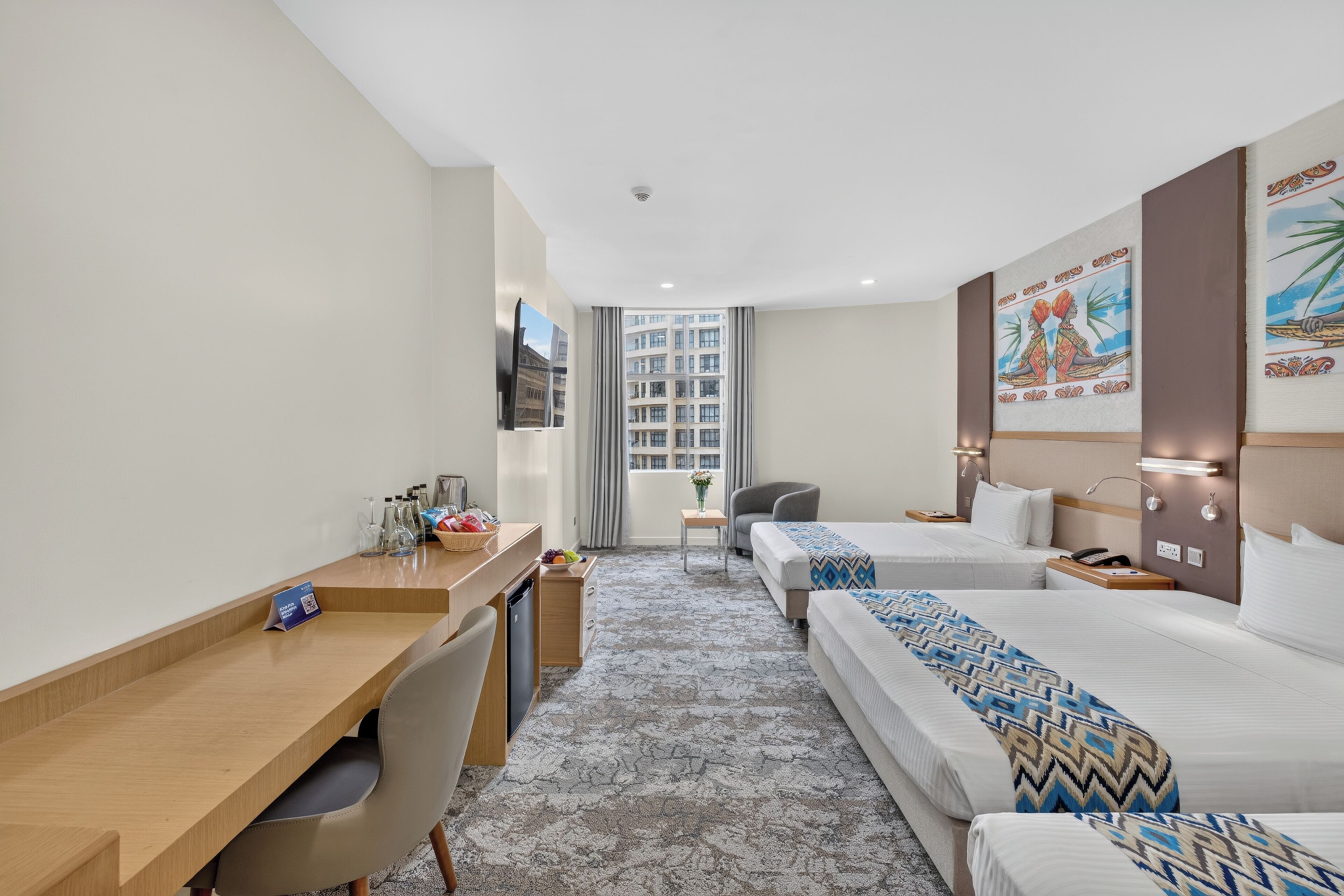 Habitación de hotel con escritorio, dos camas, una silla, una mesita y vistas a edificios a través del ventanal.