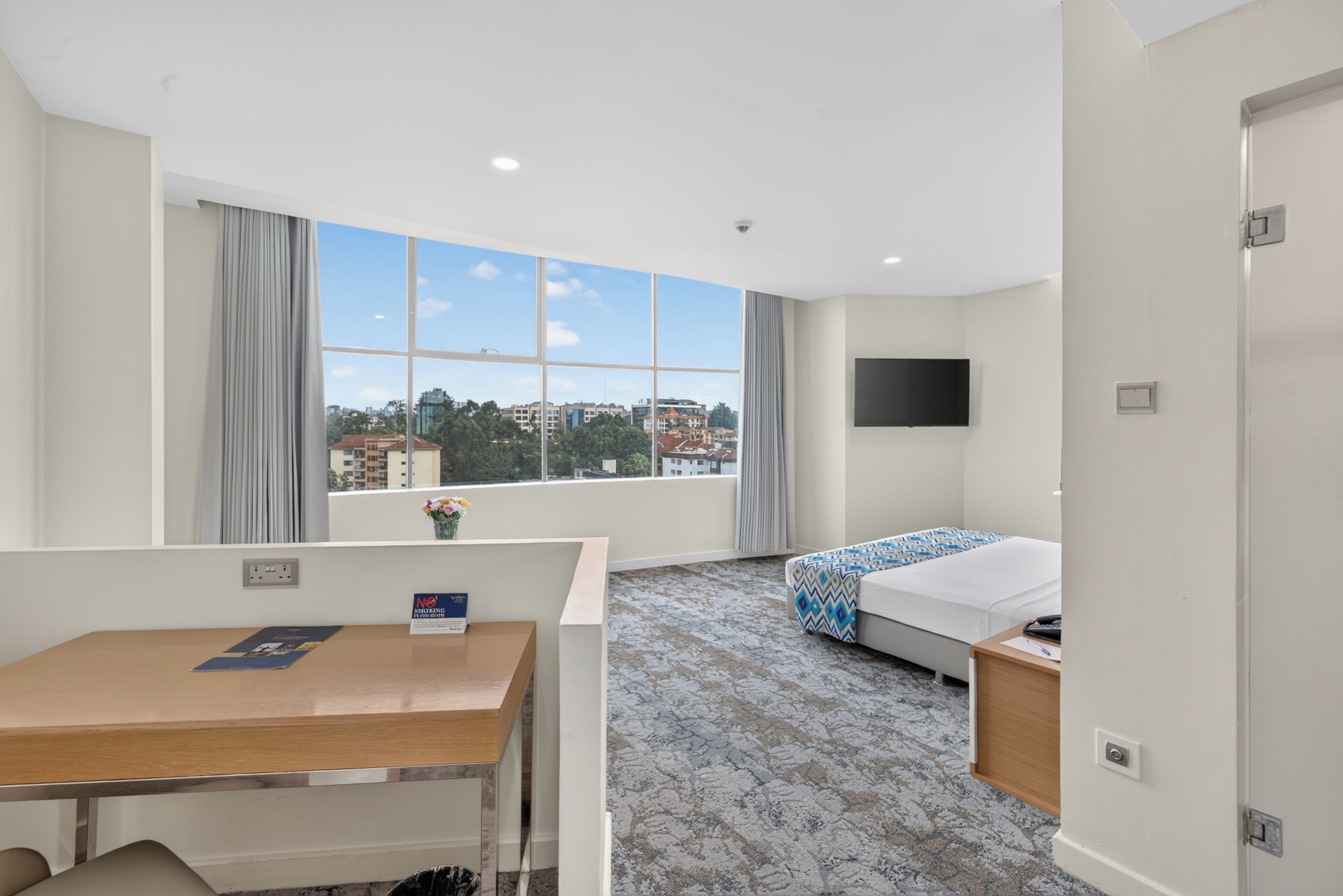 Ein Hotelzimmer mit großem Fenster, einem Bett, einem Schreibtisch und einem Fernseher.