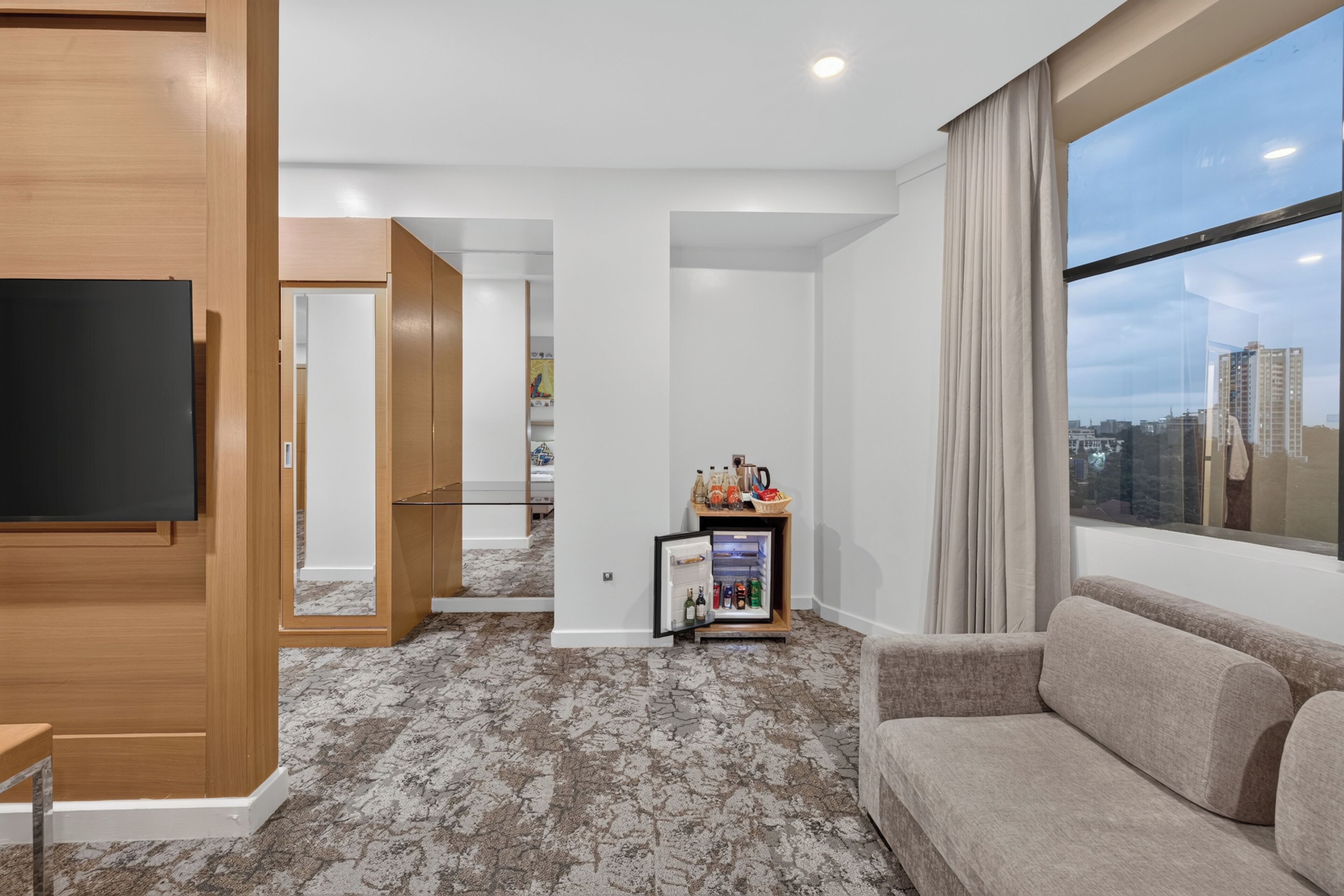 A modern hotel room with a flat-screen TV, a sofa, a mini-fridge with beverages, and a city view.