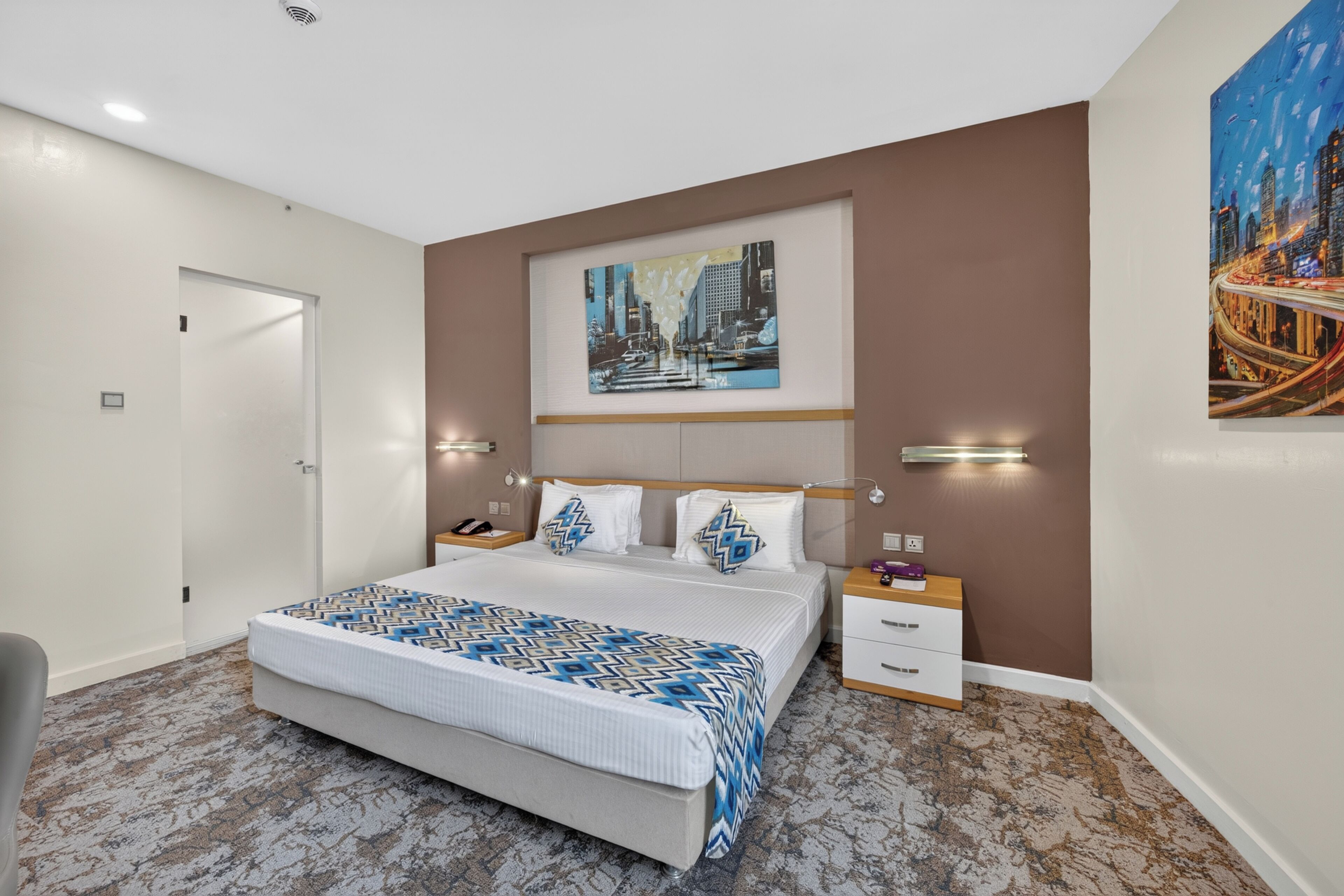 A modern hotel room with a large bed, a cityscape painting, a nightstand, and a chair.
