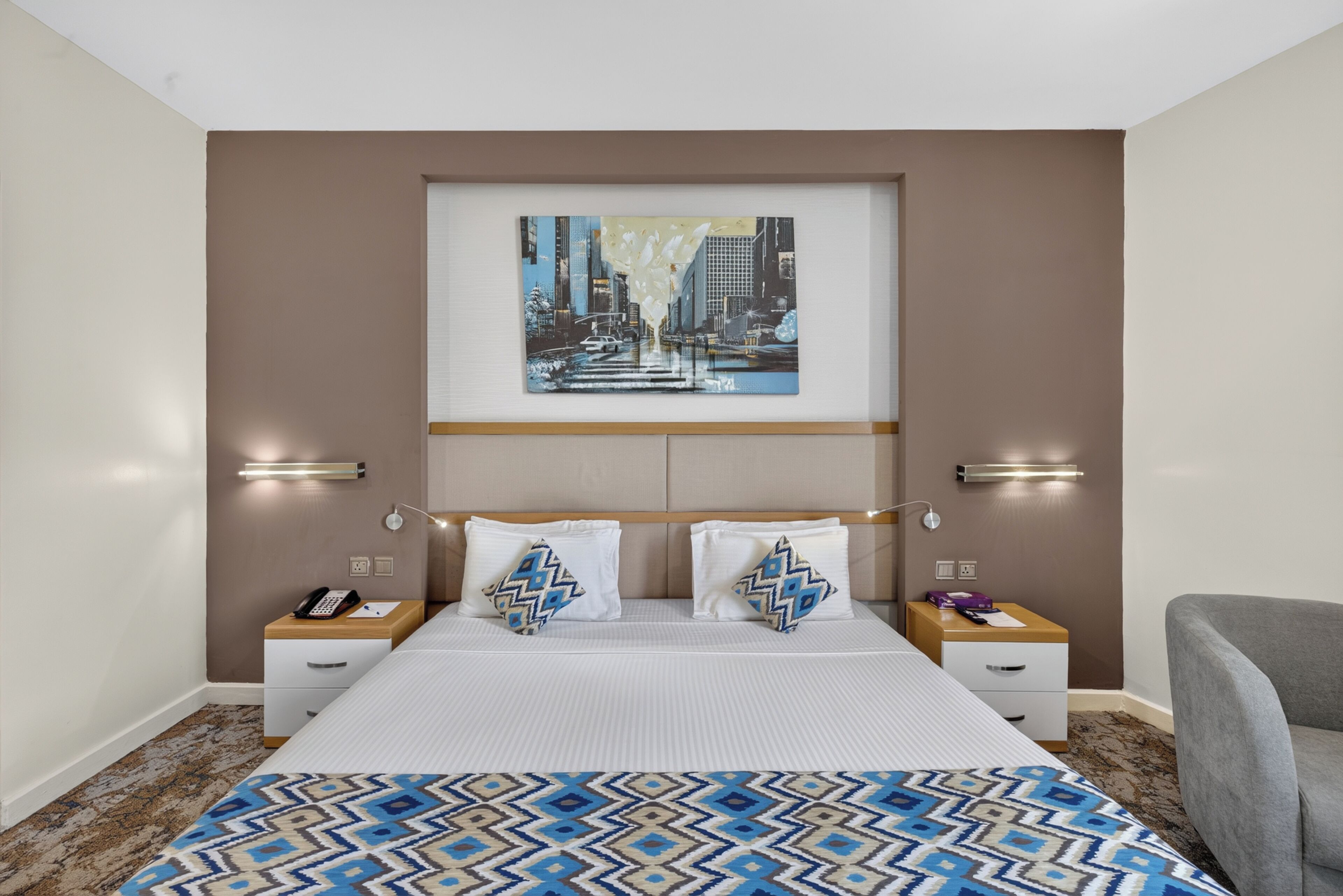 A hotel room with a large bed, two bedside tables, a nightstand, a chair, and a cityscape painting on the wall.