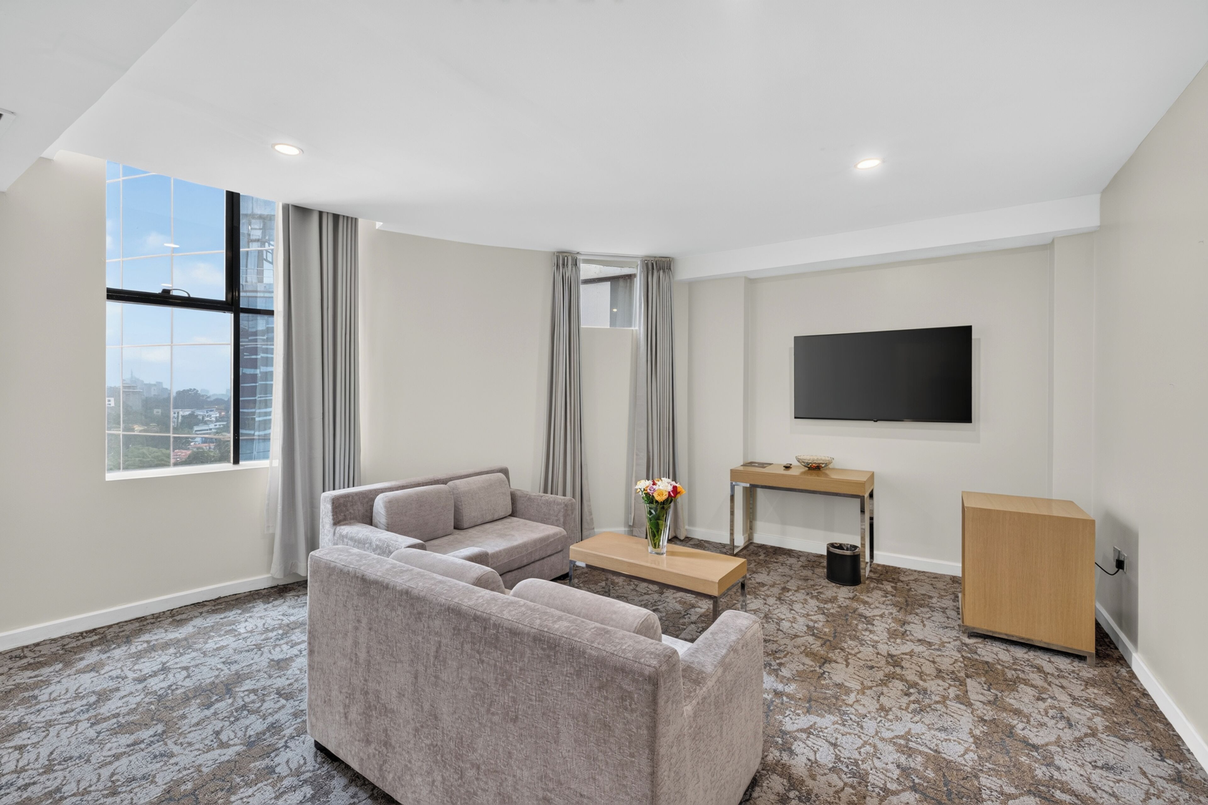 A modern hotel room with a grey sofa, a small table with a vase of flowers, a flat-screen TV mounted on the wall, and a large window with curtains.