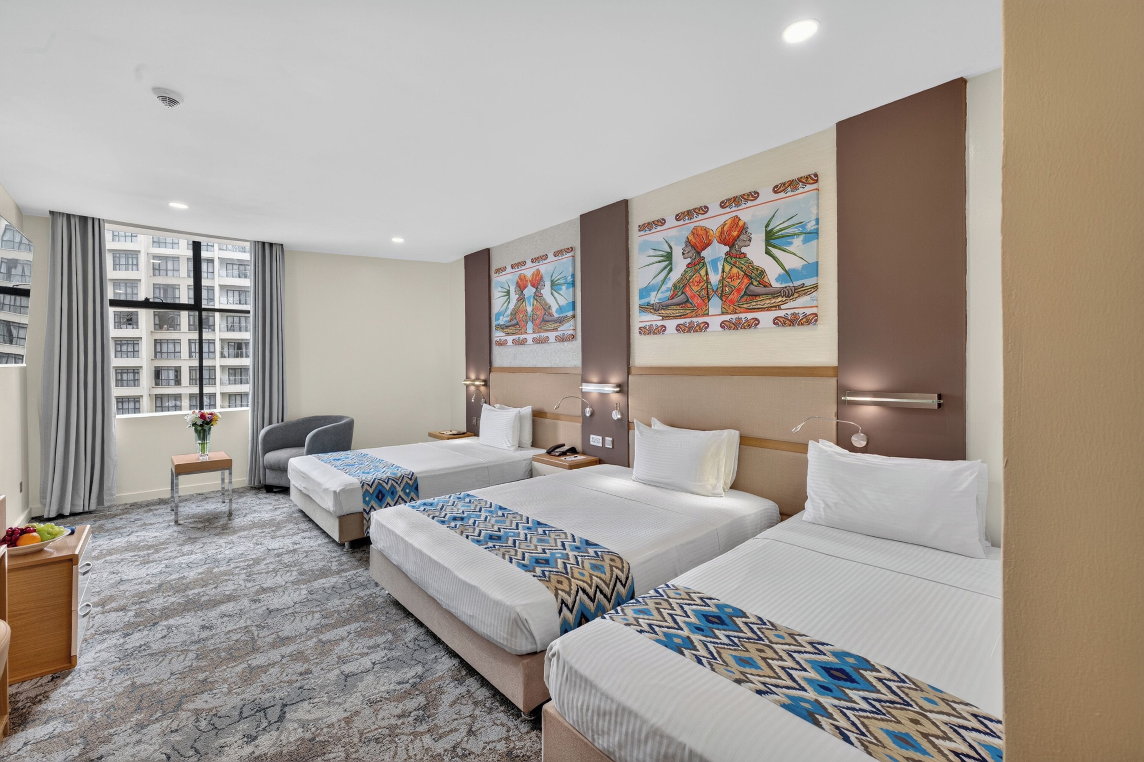 A hotel room with two beds, a desk, a chair, a window with curtains, and artwork on the wall.