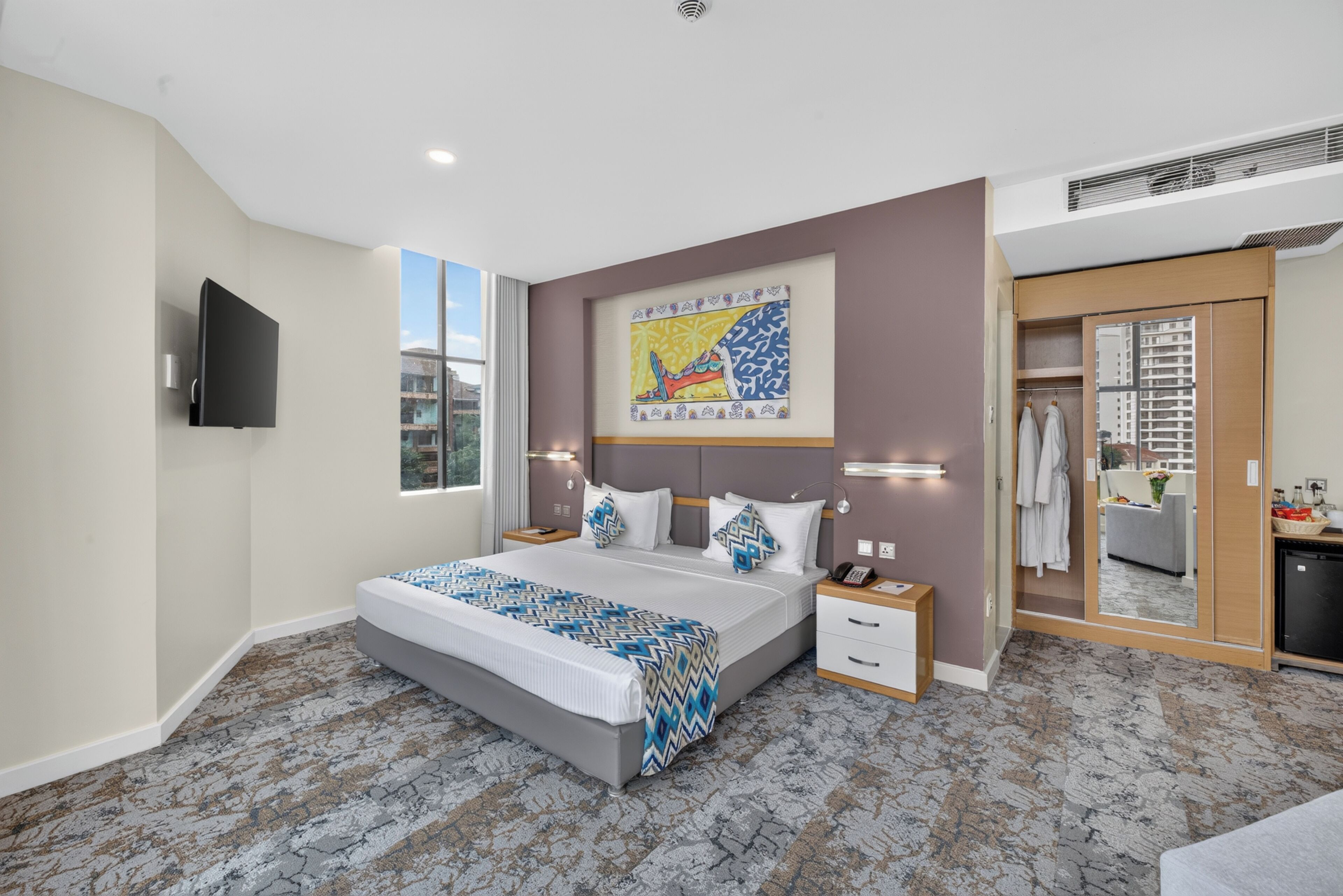 A modern hotel room with a large bed, a flat-screen TV, a painting on the wall, and a bathroom visible through an open door.