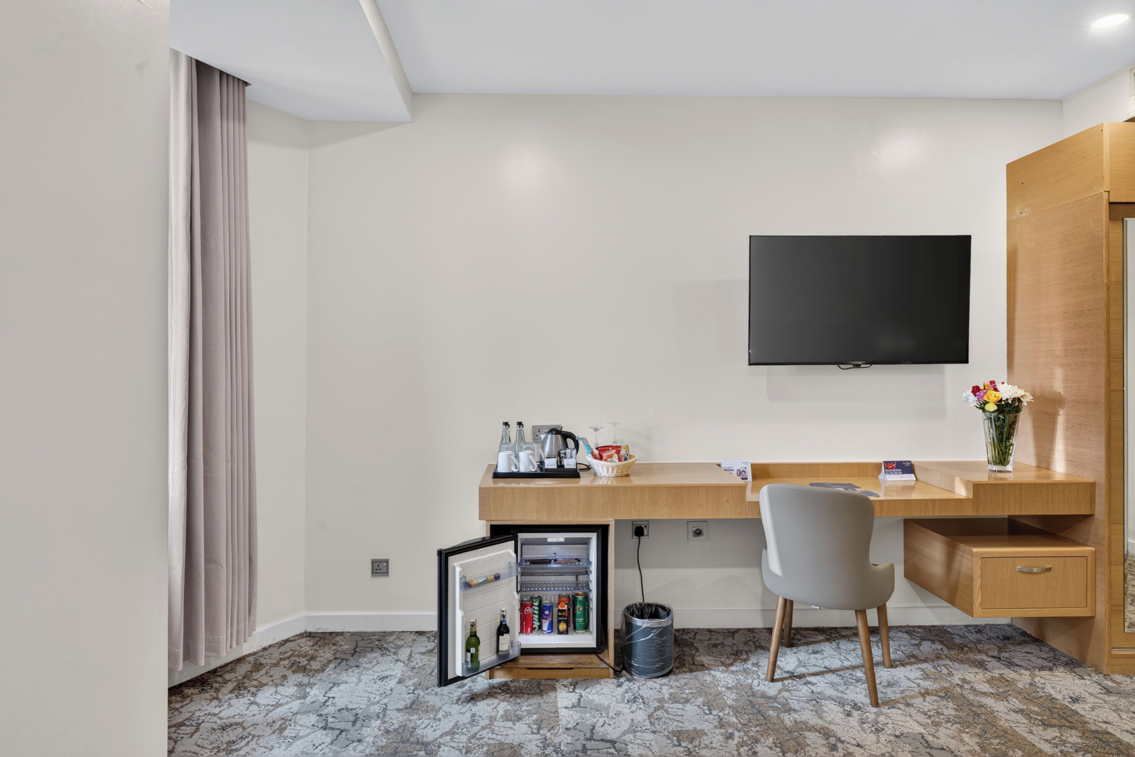 Habitación de hotel con escritorio de madera, televisor de pantalla plana, mini refrigerador, silla y cesto de basura.