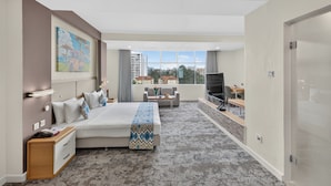 A hotel room with a bed, a TV, a desk, and a view of buildings.