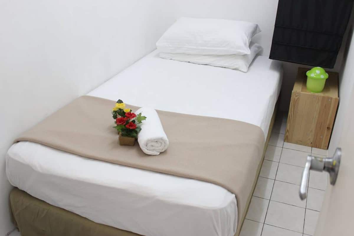 A neatly made bed with a beige blanket, a rolled white towel, and a small vase with red and yellow flowers.