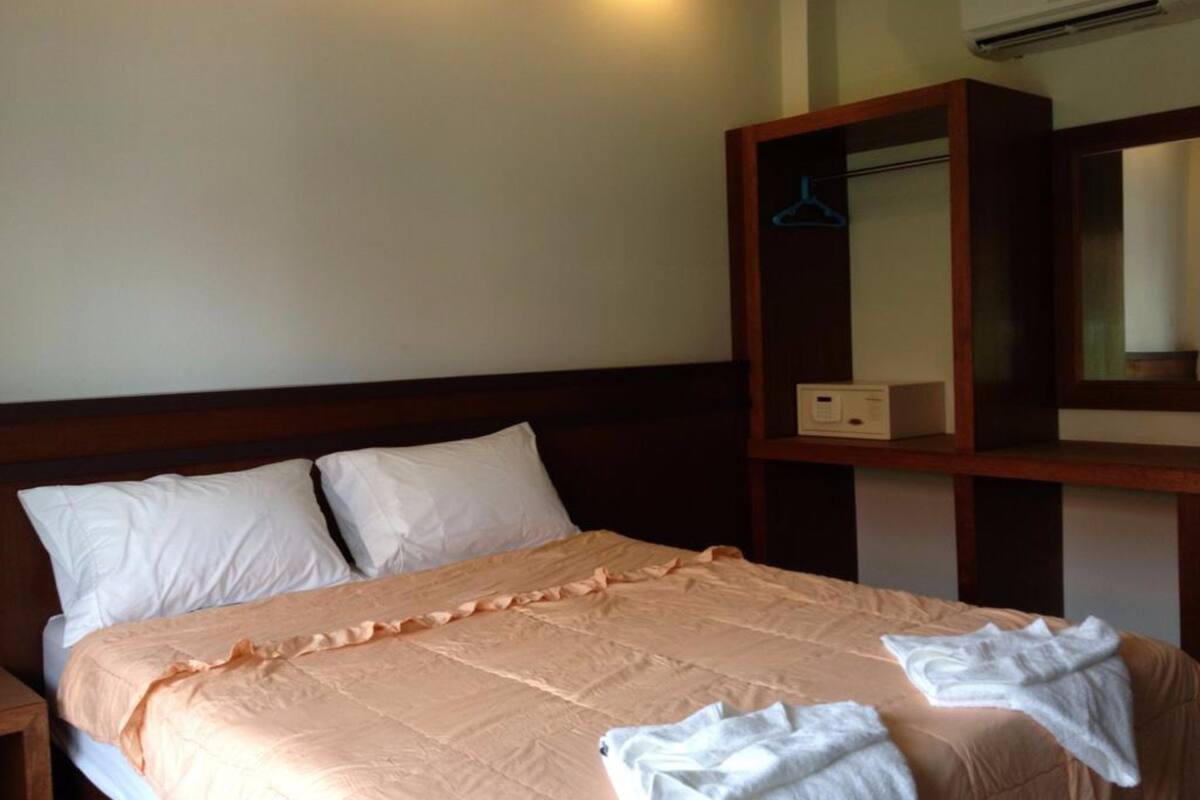 A hotel room with a bed, pillows, a bedside table, a wardrobe, and an air conditioning unit.