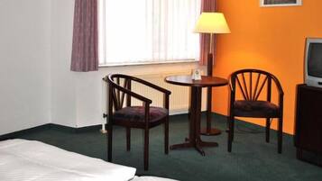 A hotel room with a bed, two chairs, a small table with a lamp, a television, and a window with curtains.