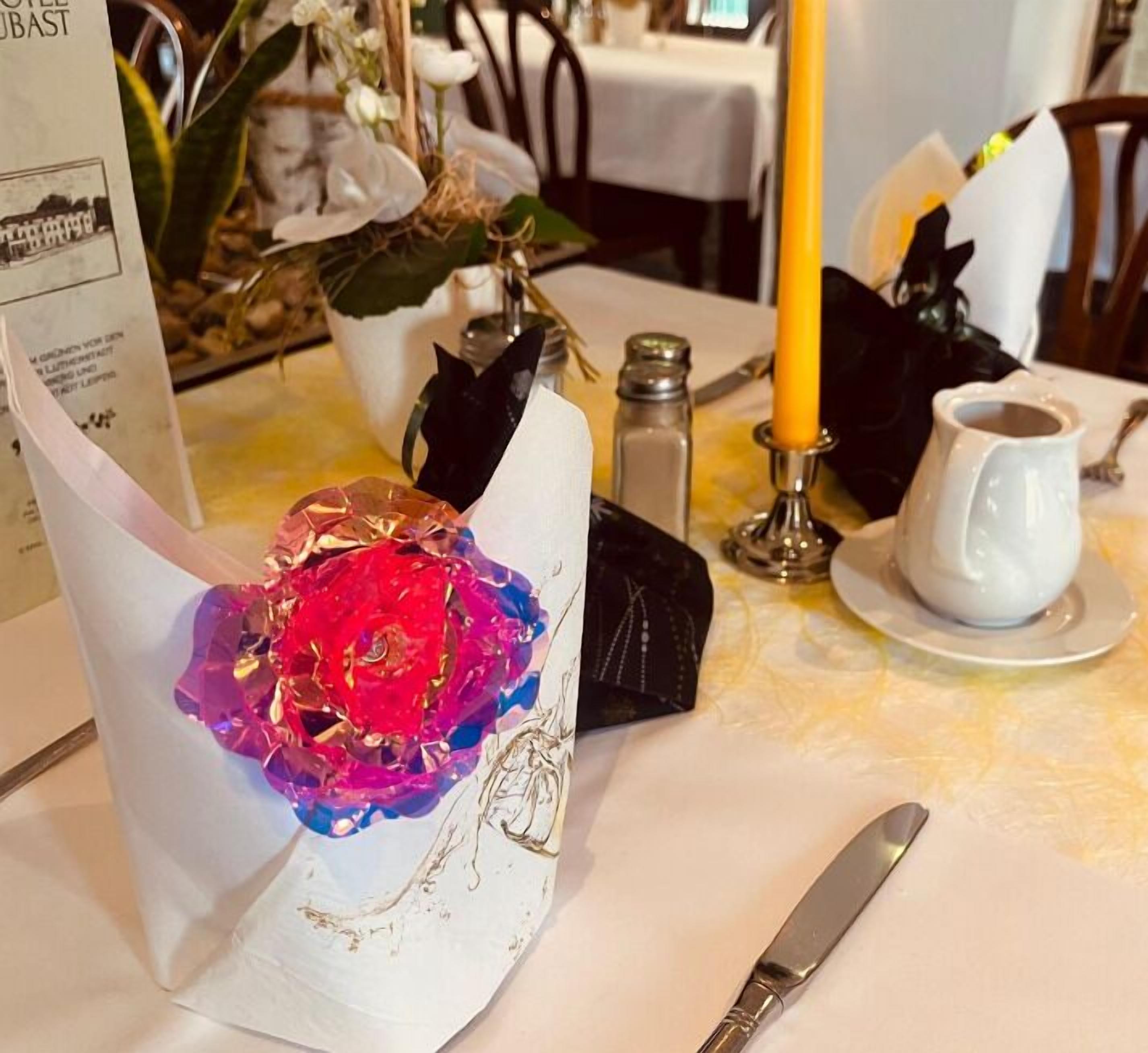 A table set with a white napkin, a glass with a colorful flower, a salt and pepper shaker, a candle, a white cup, and a knife.