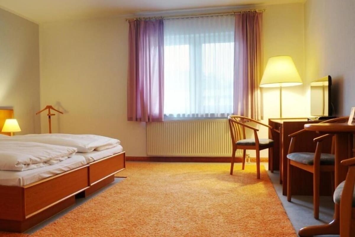A hotel room with a bed, a desk, two chairs, a lamp, and a window with curtains.