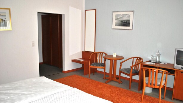 A hotel room with a bed, a desk with a chair, a television, and a framed picture on the wall. - Heidehotel Lubast (Kemberg)