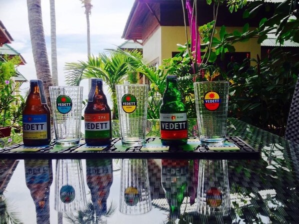 Una mesa con cuatro botellas de cerveza Vedett y cuatro vasos, reflejando la vegetación y la arquitectura circundante.