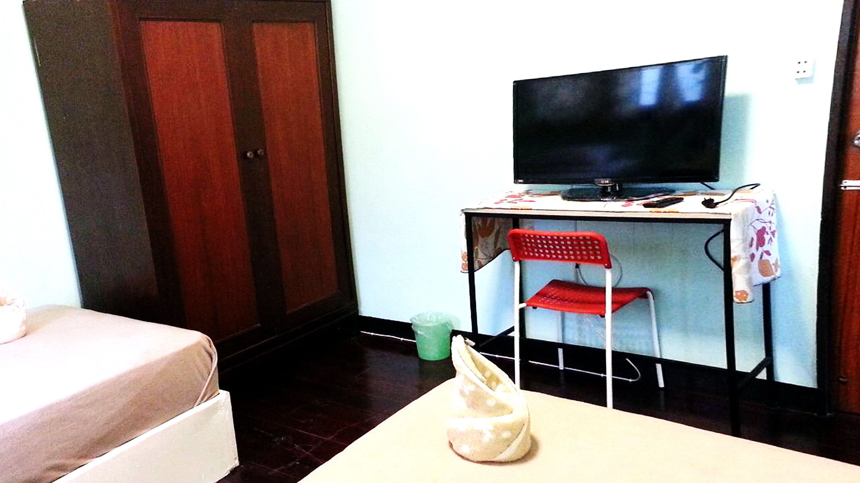 A room with a bed, a desk with a television, a red chair, and a trash bin.