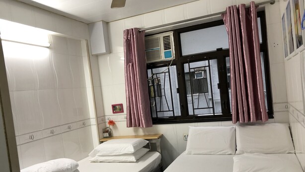A hotel room with two beds, a window with curtains, a bedside table, and an air conditioning unit. - Beverly Guest House (Kowloon)