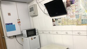 A small kitchenette with a microwave, refrigerator, and a table with a stool. There is a bulletin board with various notices and a television mounted on the wall. - Beverly Guest House (Kowloon)