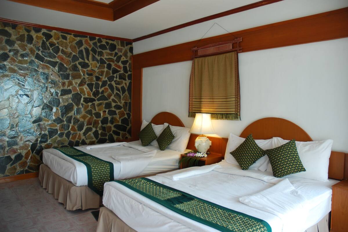 A hotel room with two beds, a stone wall, a wooden ceiling, and a window with a blind.