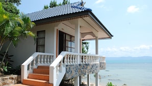 Uma casa à beira-mar com uma escadaria de pedra que dá para um alpendre, rodeada por plantas tropicais e com vista para o mar.