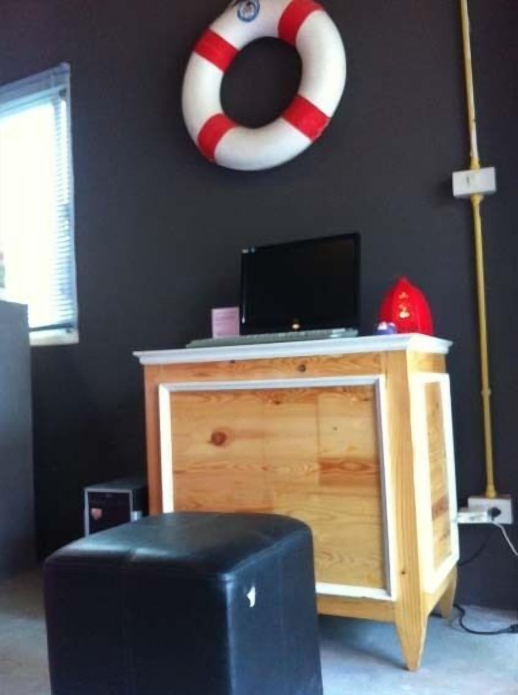 Una habitación con una pared oscura, un mueble de madera con un monitor negro, un casco rojo y un taburete negro.