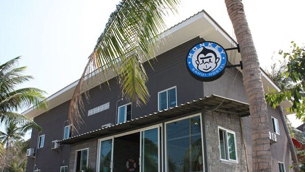 A building with a large glass facade, a palm tree in front, and a logo on the side. - Monkey Samui Hostel (Koh Samui)
