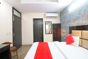A hotel room with a bed, a desk, a chair, an air conditioning unit, and a door. - Westend Holiday Home (New Delhi)