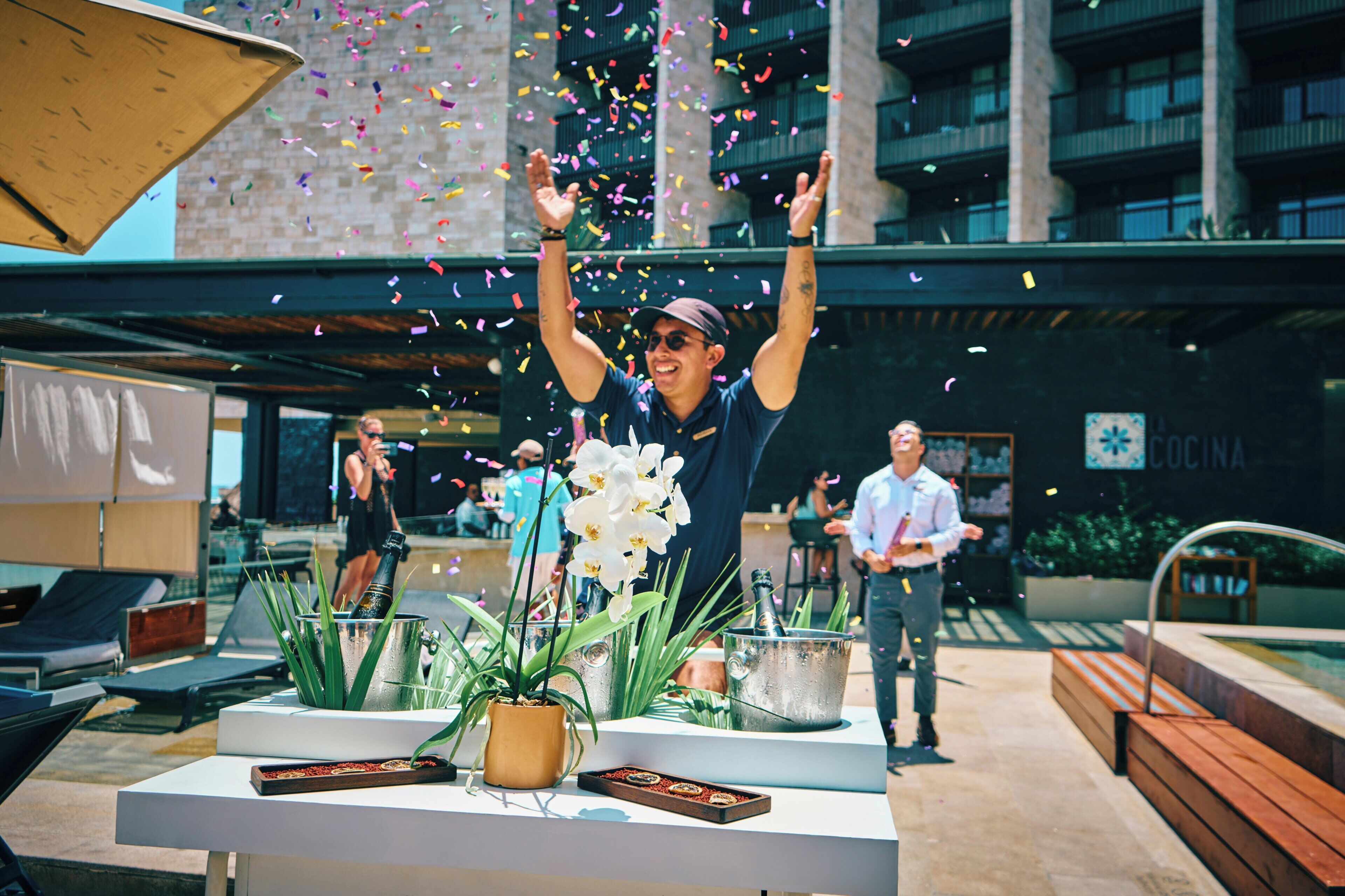 A person celebrating with confetti, a table with a potted plant, and a building with balconies in the background.