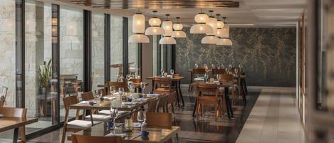 A modern restaurant with wooden tables and chairs, hanging pendant lights, and large windows.