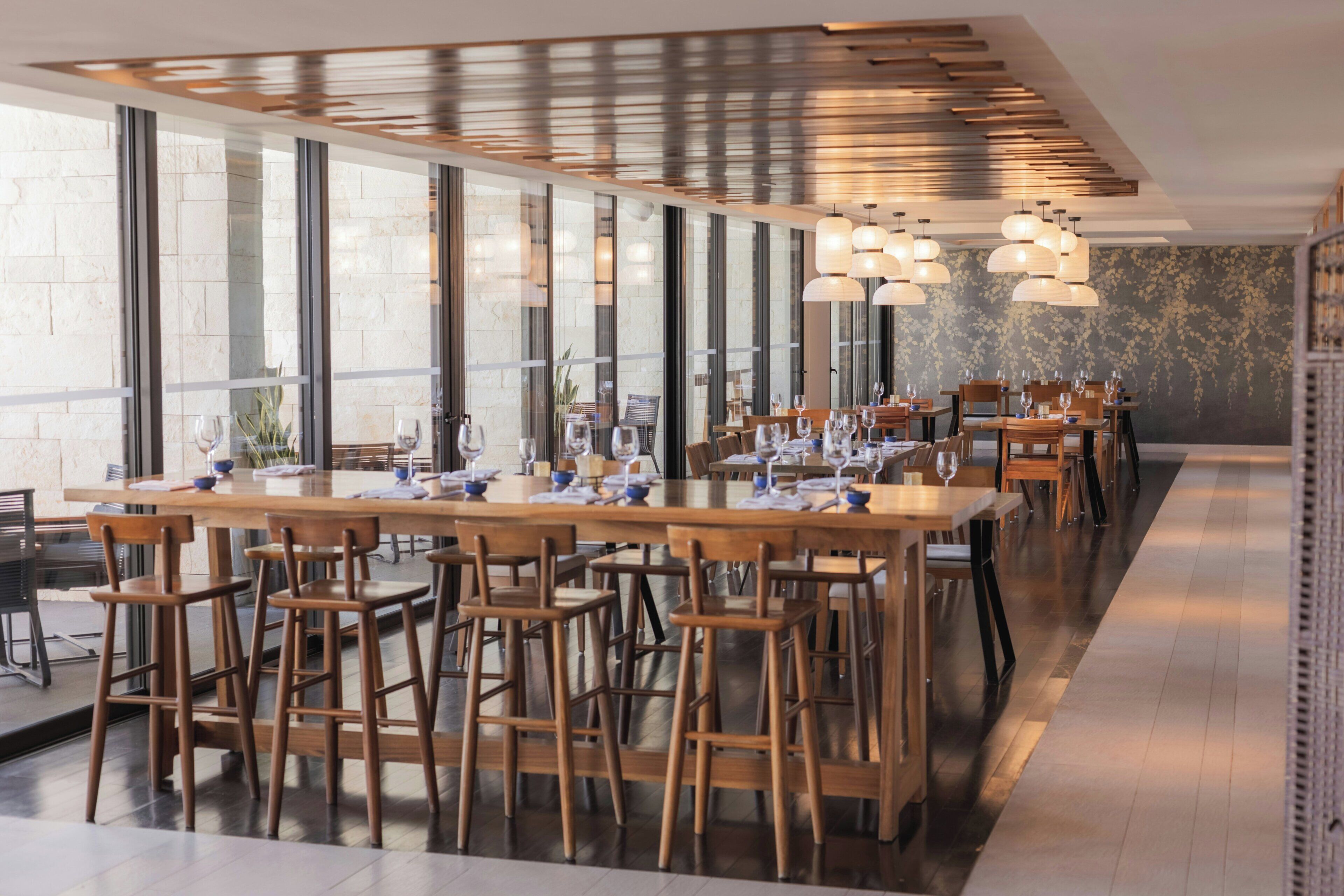 Un restaurant moderne doté d’une longue table à manger, de nombreuses chaises et de luminaires suspendus.