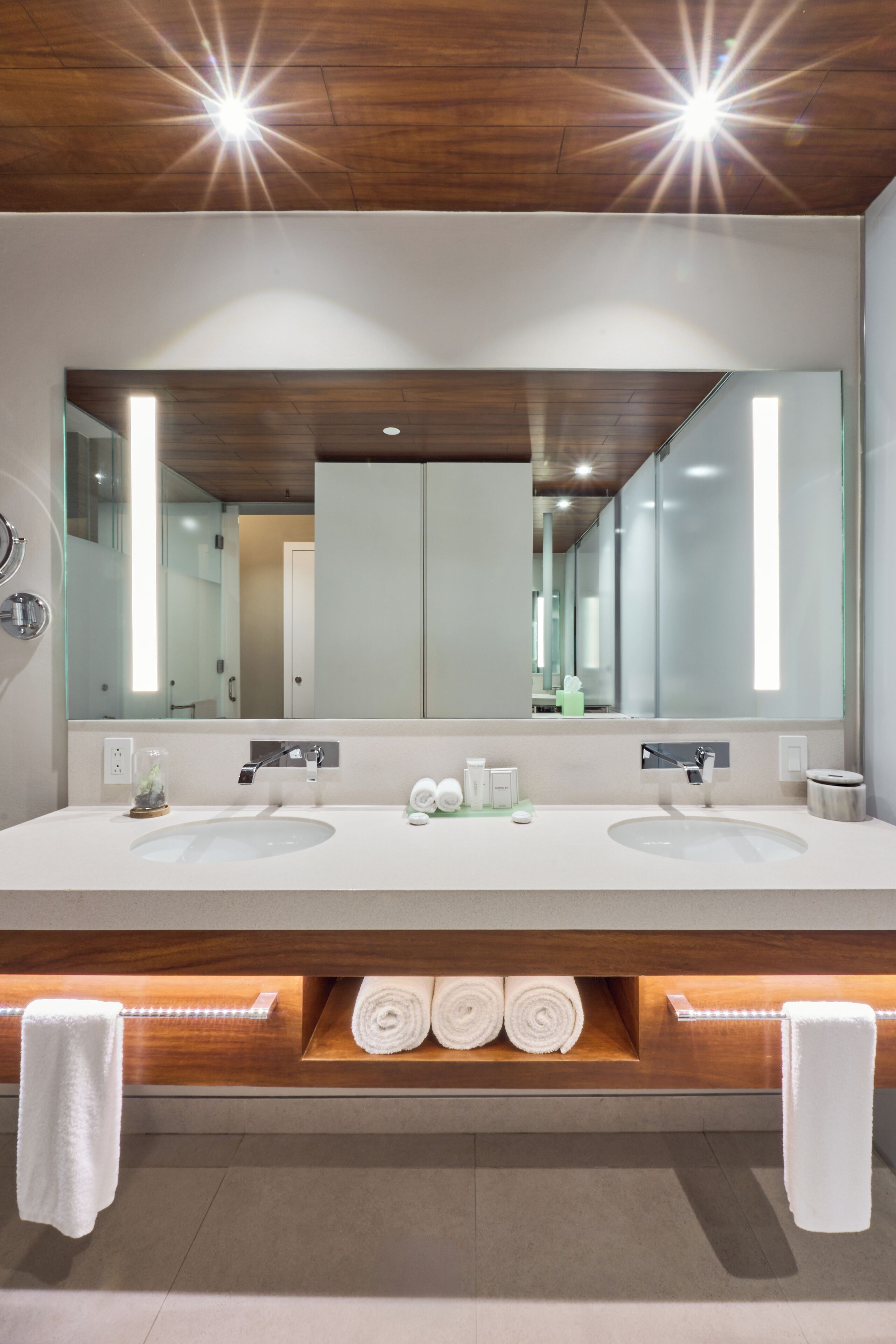 Une salle de bain moderne avec deux vasques, un grand miroir et des étagères en bois où sont posées des serviettes.