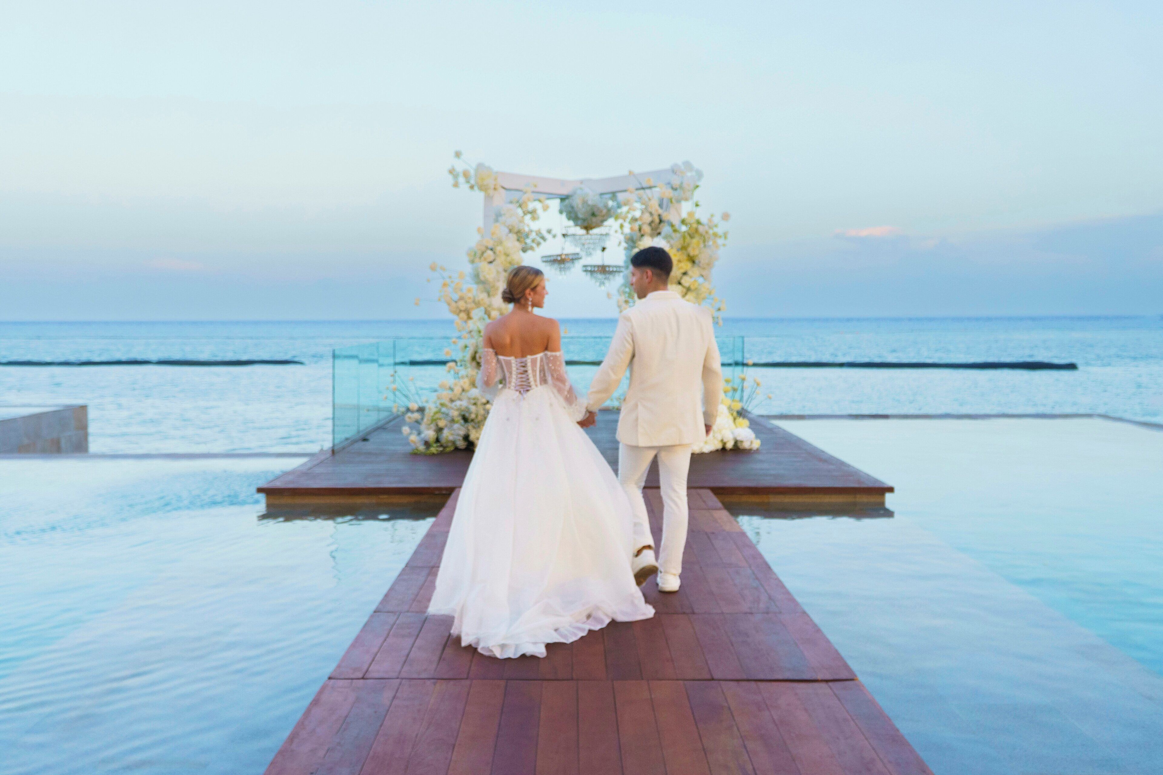 A couple walking on a wooden deck towards a floral arch over water.