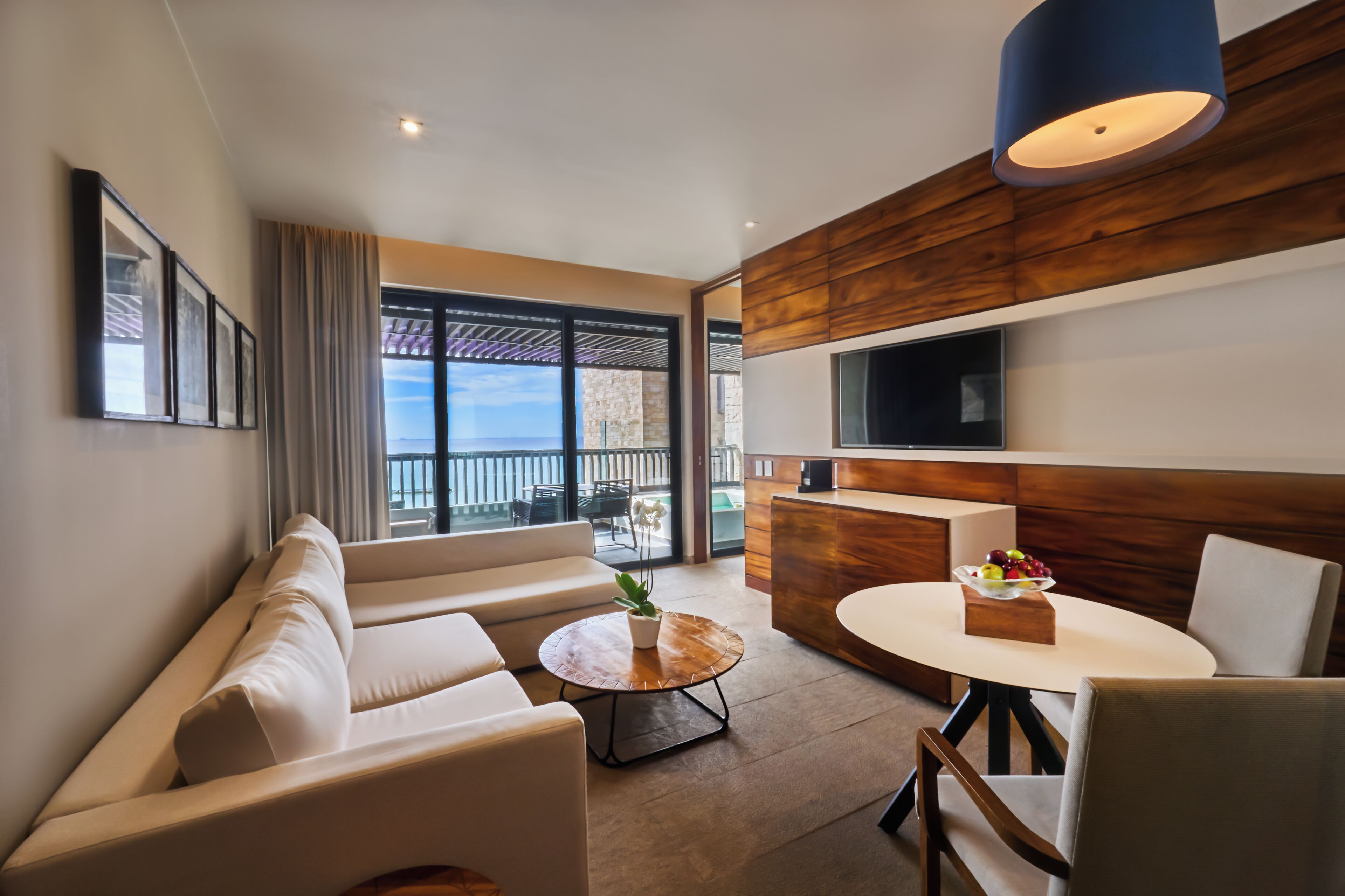 Una sala de estar moderna con un sofá, una mesa redonda de centro con una planta, una mesa de comedor con sillas, un televisor montado en la pared y vistas al mar a través de amplios ventanales.