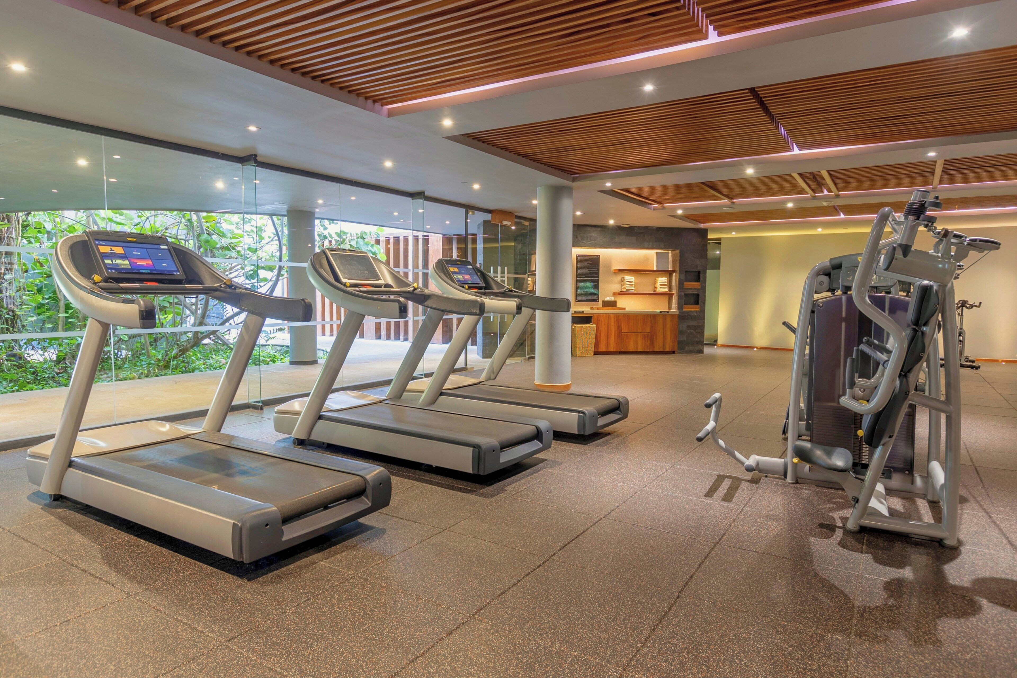 A modern gym with treadmills, a weight machine, and a reception area with a view of greenery.