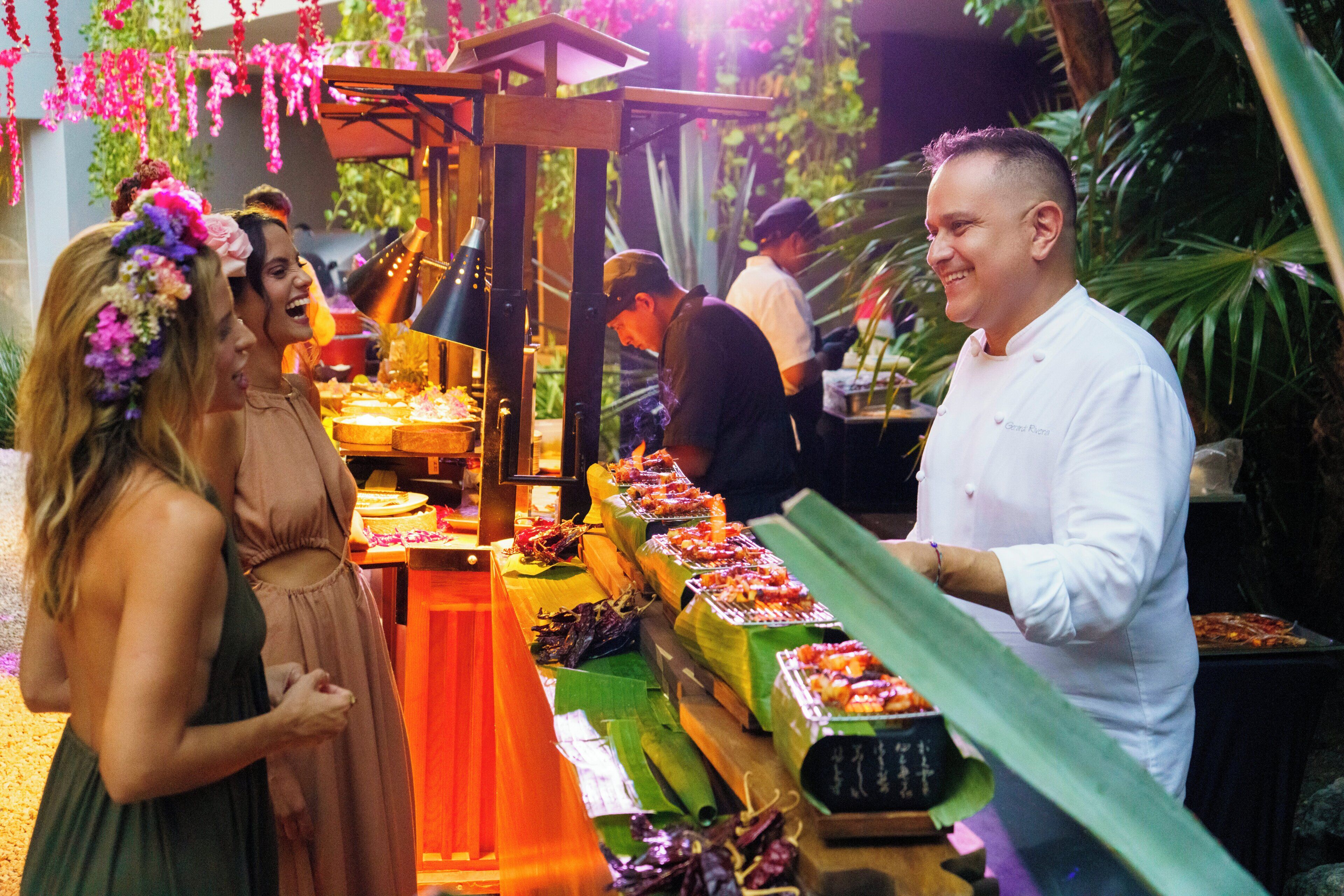 A food stall with various dishes displayed on a table, surrounded by people.