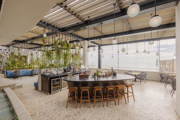 A modern bar area with a central island, bar stools, hanging plants, and a view of the sky through large windows. - Devossion By Live Aqua Playa del Carmen All Inclusive - Adults Only (Playa del Carmen)