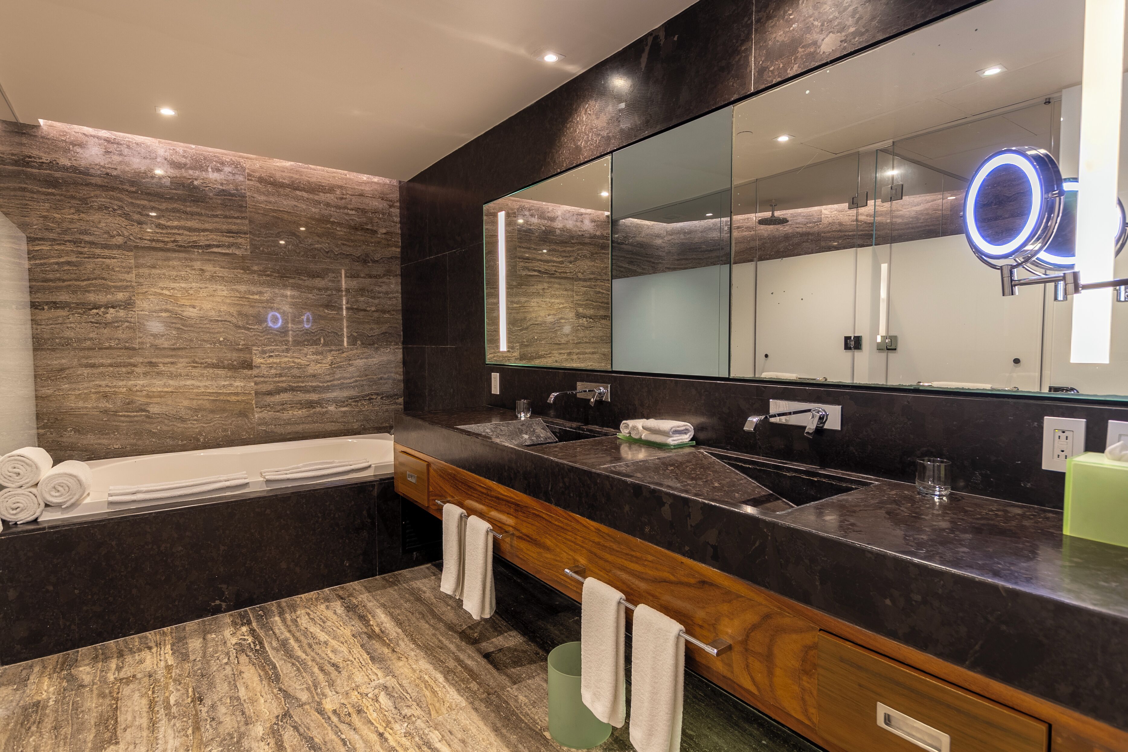 A modern bathroom with a large mirror, dual sinks, a bathtub, and a vanity with a wooden cabinet.