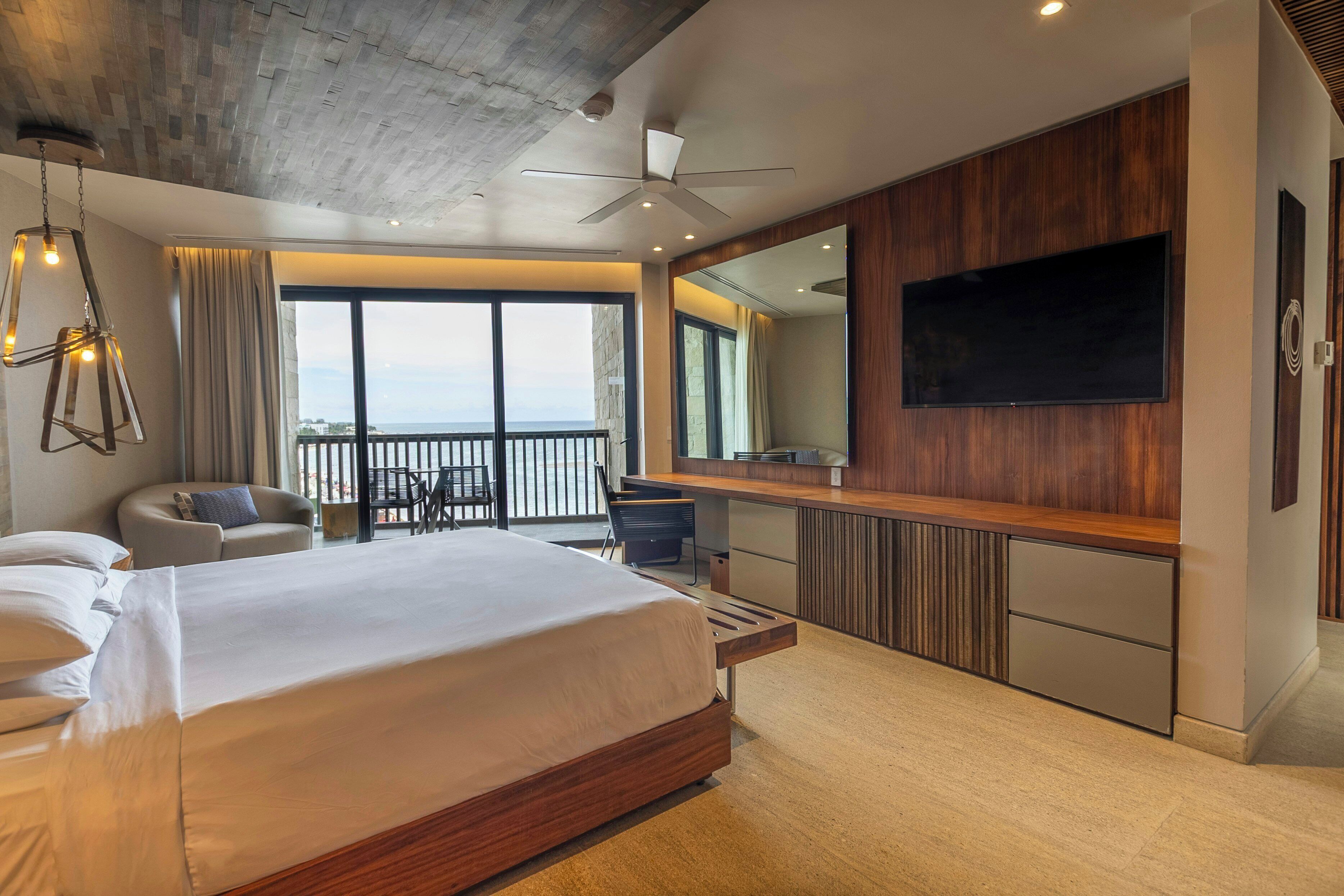 A modern hotel room with a large bed, a desk, a TV, and a balcony with a view.