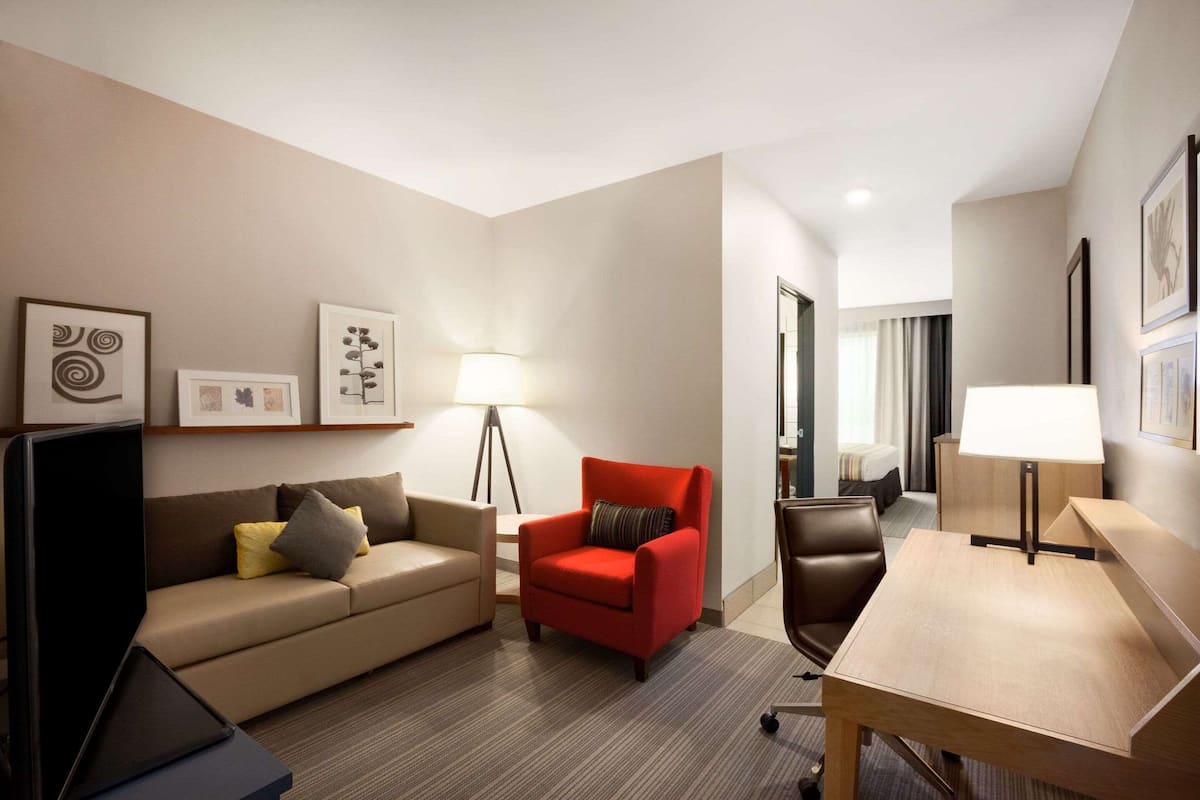 A hotel room with a sofa, a red armchair, a desk, a lamp, and framed artwork on the wall.