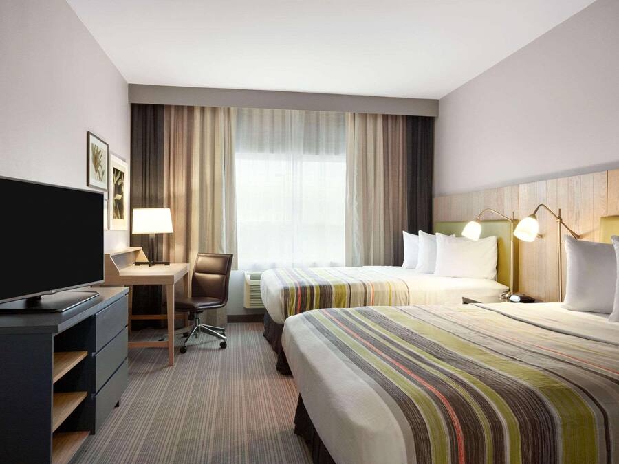 Habitación de hotel con televisor, escritorio, dos camas, una silla y un ventanal grande con cortinas.