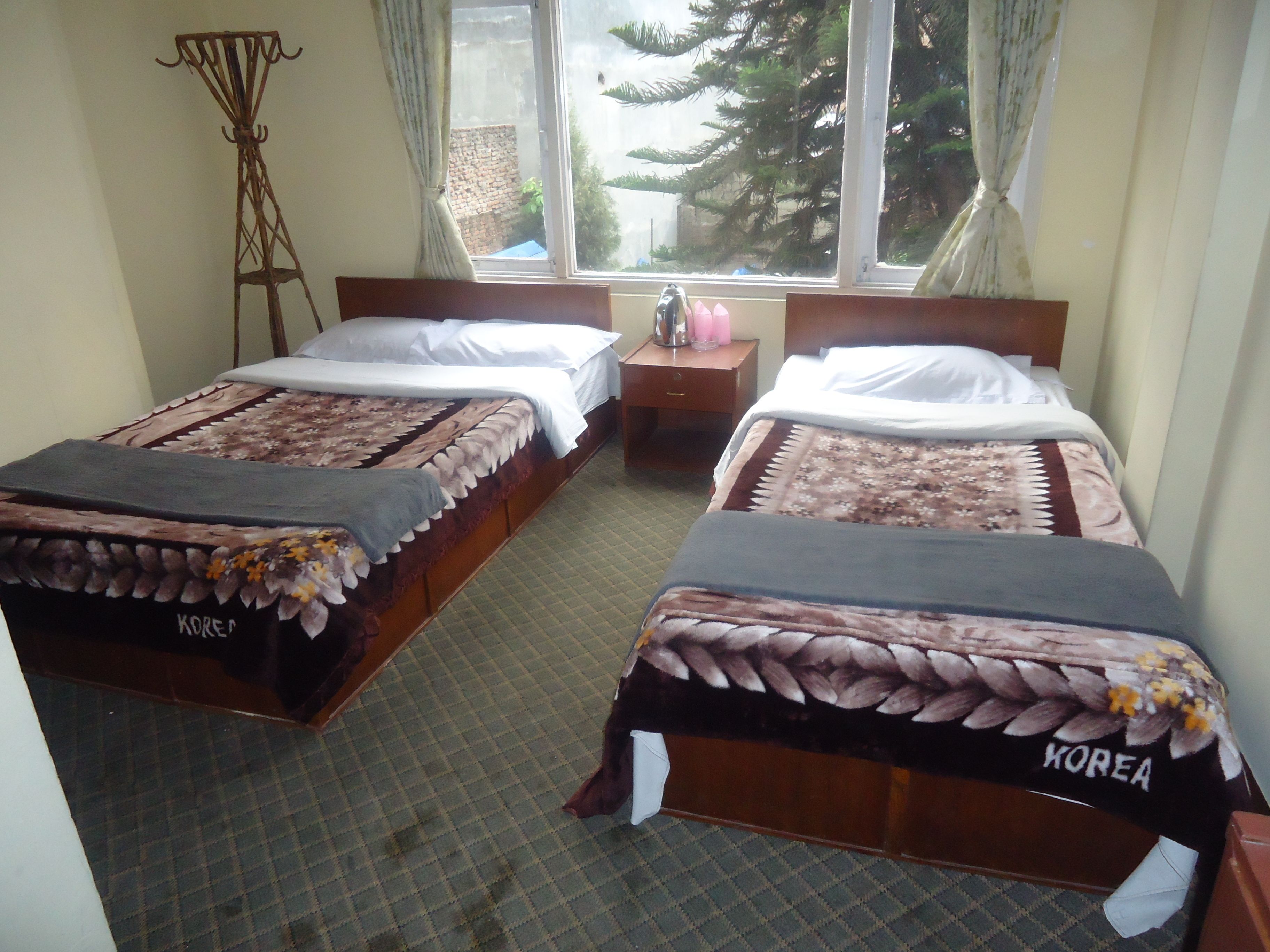 A room with two beds, each with a floral bedspread and a blanket labeled "KOREA". There is a nightstand with a lamp and a vase of flowers between the beds.