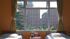 A room with two beds, a window with curtains, a small table with a kettle and pink bottles, and a view of brick buildings and greenery outside. - Hotel Discovery Inn (Kathmandu)